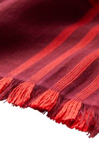 Couverture tissée rouge foncé et violette avec des franges orange, présentant des rayures alternées de différentes textures.