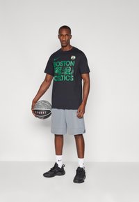Czarny t-shirt Nike z krótkim rękawem z napisem "BOSTON CELTICS" w kolorze zielonym, szare szorty, czarne sneakersy, trzymając piłkę koszykową. Prosty, sportowy design.