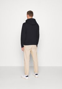 Felpa nera in cotone con cappuccio regolabile, abbinata a pantaloni beige e sneaker bianche. L'outfit è visto da dietro su uno sfondo bianco.