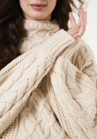 Beige kabelgebreide trui met een hoge hals en oversized mouwen. Gelaagde patronen creëren diepte; ribgebreide boorden voegen detail toe. Zachte stof.