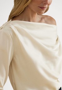 Krém színű, vállakat szabadon hagyó blúz drapírozott elülső résszel, selymes szatén textúrával és hosszú ujjakkal, amelyek finoman összegyűrt részleteket mutatnak a vállnál.