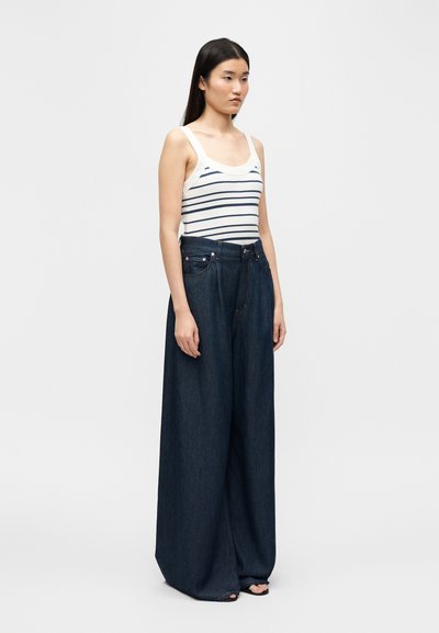 Woman wearing a white tank top with navy stripes and high-waisted, wide-leg dark blue jeans standing against a plain light background.