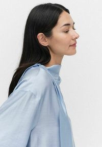 Vaaleansininen, satiinimaineinen paita, jossa on korkea kaula-aukko ja valuva hihansuut. Ominaisuuksiin kuuluu pehmeä laskostus ja ainutlaatuinen solmuseikka olkapäällä.