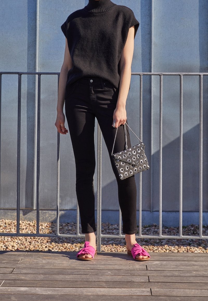 Black sleeveless turtleneck sweater, black fitted jeans, bright pink slip-on sandals with bows, and a small patterned black and white handbag.