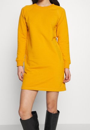 Nainen pukeutuneena pitkähihainen sinappikeltainen neulemekko ja mustat polven yläpuolelle ulottuvat saappaat, seisomassa yksinkertaista valkoista taustaa vasten.