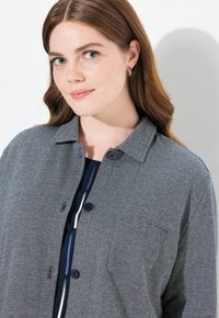 Chaqueta de camisa en espiga gris con cuello, dos botones y un bolsillo en el pecho, llevada sobre una parte superior oscura con finas rayas blancas y azules.