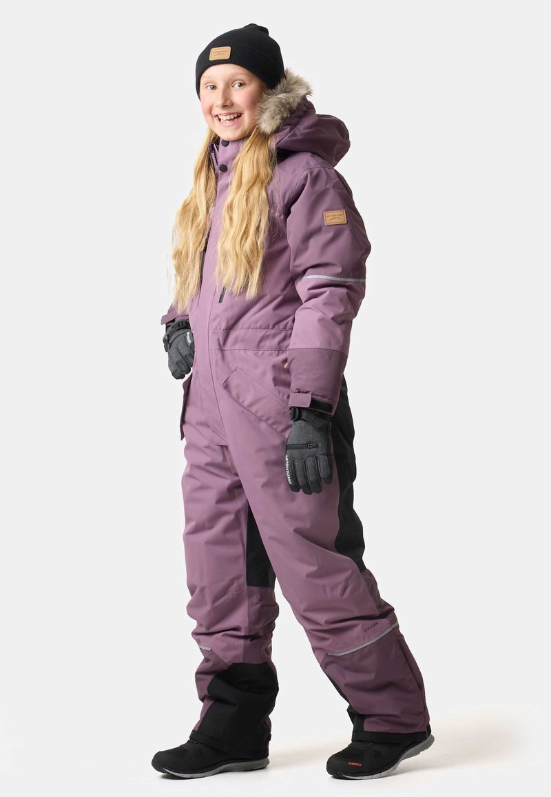 Lila isolierter Schneanzug mit einer mit Fell gefütterten Kapuze, schwarzen Akzenten, reflektierenden Streifen und schwarzen Handschuhen, getragen mit schwarzen Winterstiefeln.