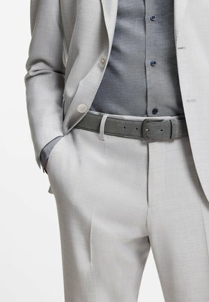 Homme portant un costume gris clair avec une chemise gris foncé rentrée dans le pantalon et une ceinture grise assortie, main dans la poche.