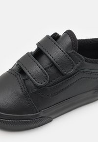 Chaussure en cuir noir avec deux bandes Velcro sur la languette, surface texturée, bout arrondi et semelle en caoutchouc avec motif de grip.