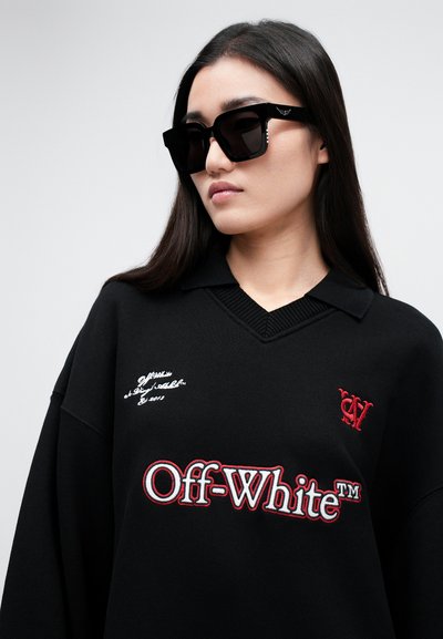 Jauna sieviete, kas valkā pārmēru lielas melnas saulesbrilles un melnu Off-White™ džemperi ar baltajiem un sarkanajiem logotipiem priekšpusē.