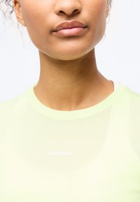 Κοντινό πλάνο του κάτω μέρους του προσώπου και του λαιμού ενός ατόμου που φοράει ανοιχτό κίτρινο πλεκτό μπλουζάκι με ορατό λευκό λογότυπο "Salomon".