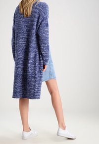 Lång, kraftig stickad cardigan i blått med ett texturerat mönster. Kombinerad med en ljusblå jeanskirt och vita sneakers.
