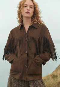 Brown suede jacket featuring fringe accents, button closure, and two front pockets. The design has a relaxed fit with a collar and textured surface.