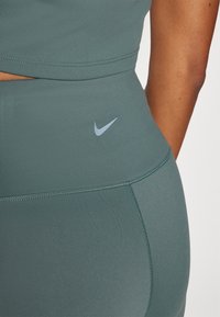 Leggings atletici verdi realizzati in un materiale liscio e elastico. Presentano un logo Nike azzurro chiaro in vita e dettagli di cucitura minimi.