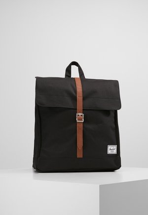 Sac à dos carré noir avec une sangle de fermeture à boucle en cuir marron, une poignée supérieure et une étiquette de marque blanche rectangulaire en bas à droite.