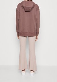 Felpa marrone con cappuccio e vestibilità comoda, abbinata a pantaloni svasati a coste chiari. Scarpe bianche e sfondo neutro completano il look.