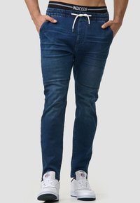 Pantalones jogger de mezclilla en azul oscuro, con cintura ajustable, bolsillos laterales y detalles acanalados para mayor textura. Combinados con zapatillas blancas.