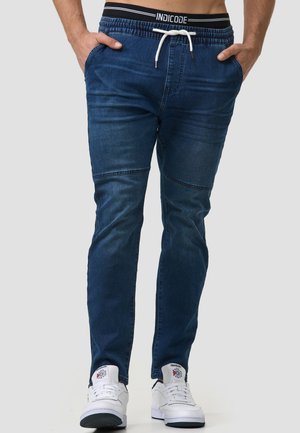 Pantalones jogger de mezclilla en azul oscuro, con cintura ajustable, bolsillos laterales y detalles acanalados para mayor textura. Combinados con zapatillas blancas.