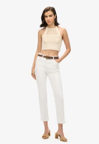 Crèmekleurige gehaakte haltertop met uitsnijdingen, gecombineerd met witte rechte broek en een bruine leren riem. Beige blokhak sandalen.