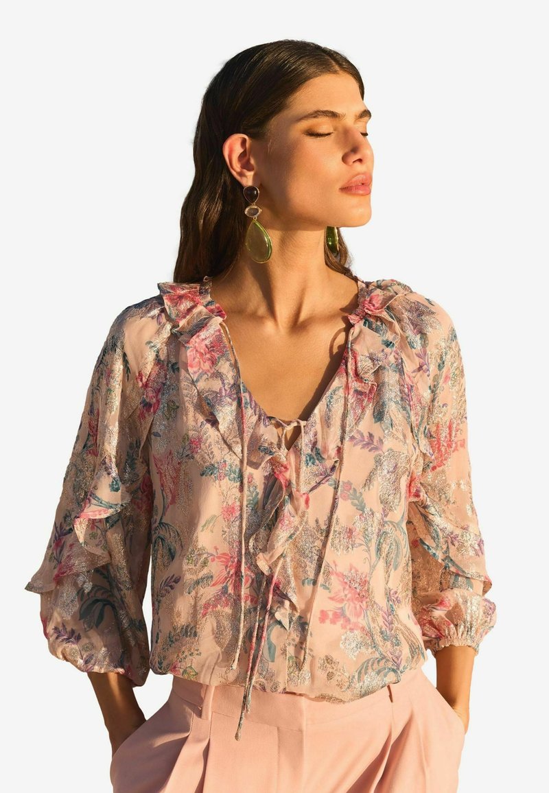 Floral blouse in soft pink chiffon with ruffled neckline and sleeves. Features intricate patterns and a tie closure at the front.
