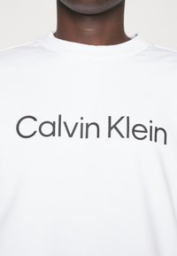 Witte katoenen T-shirt met een groot zwart "Calvin Klein" logo op de borst. Ronde hals, korte mouwen, gladde textuur.
