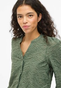 Blusa verde texturizada con un patrón ondulado, que cuenta con un cierre de botones en el frente y un cuello. Tela ligera y semitransparente con mangas largas.