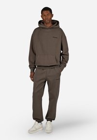 Hnedá mikina s kapucňou, predným vreckom a logom, v kombinácii s nohavicami na cvičenie v rovnakom štýle. Hladká, jemná tkanina s rebrovanými manžetami a pásom. Biely tenisky.