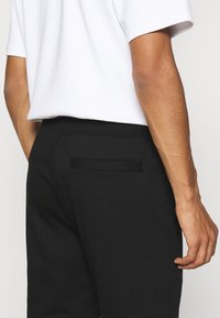 Schwarze Jogginghose mit glatter Textur, ausgestattet mit einer Gesäßtasche und einem elastischen Bund, kombiniert mit einem schlichten weißen T-Shirt.