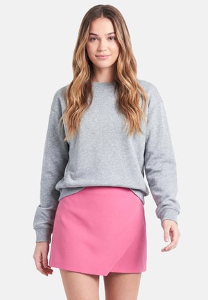 Junge Frau mit langem welligem Haar, die einen hellgrauen Sweatshirt und einen leuchtend pinkfarbenen asymmetrischen Minirock trägt, steht vor einem einfarbigen Hintergrund.