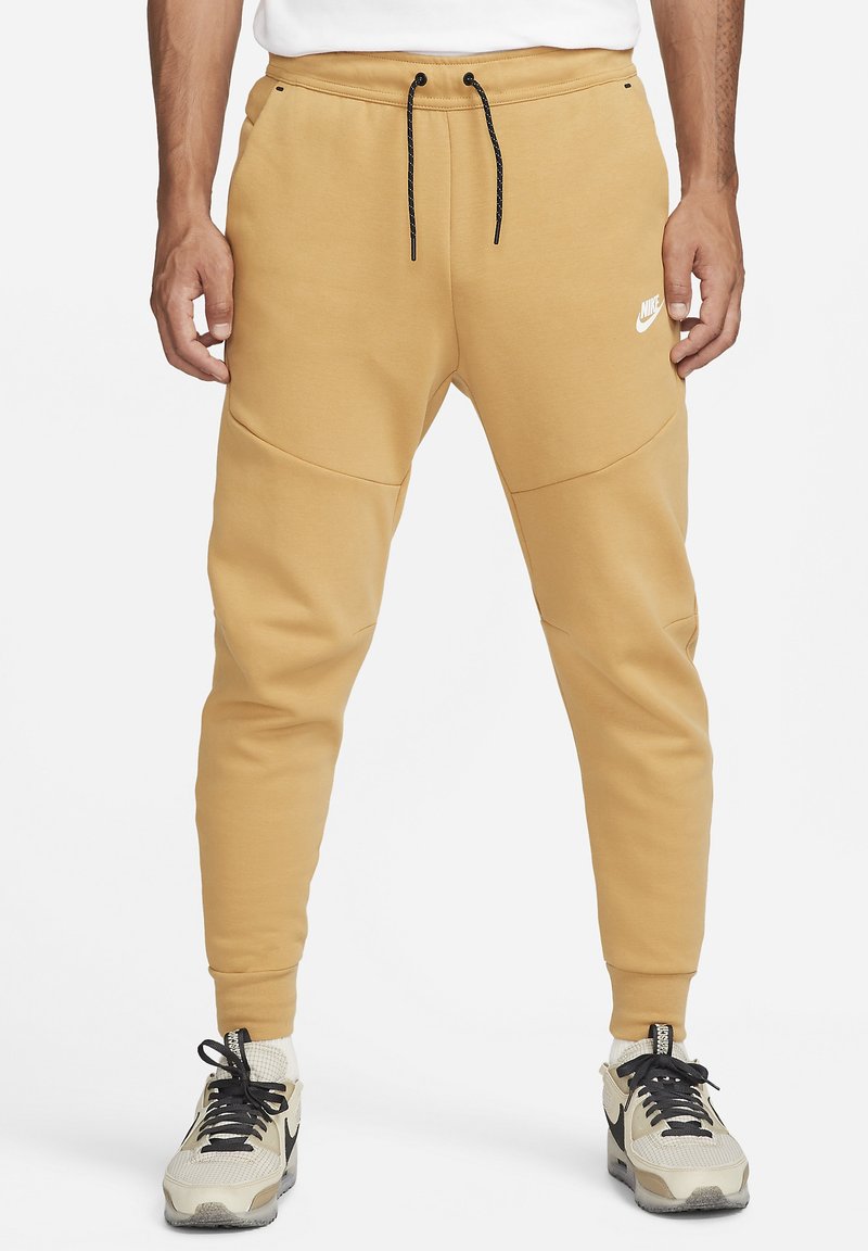 Nike joggers in light brown, featuring a drawstring waistband, fitted cuffs, and side seam accents. Pairing with beige and black sneakers.