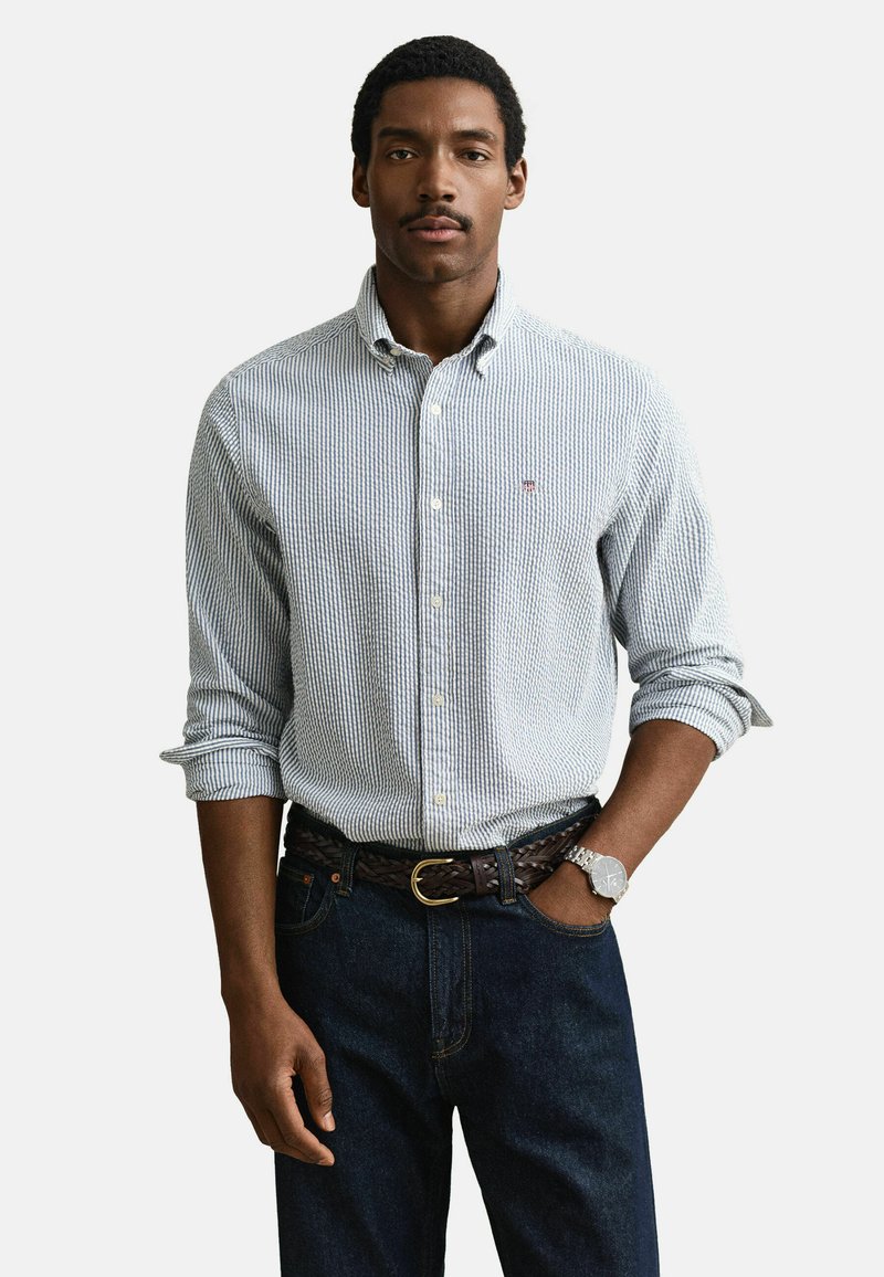 Hombre vestido con camisa de rayas azul claro de botones, vaqueros oscuros, cinturón trenzado marrón, reloj de pulsera plateado, de pie con una mano en el bolsillo.