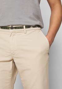 Beige trousers with a relaxed fit, featuring a camouflage patterned belt and a button closure, made from lightweight fabric.