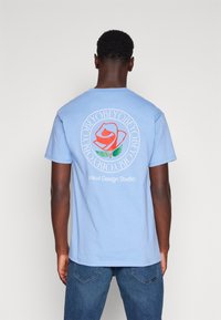 Svetle modré bavlněné tričko s krátkým rukávem a kulatým výstřihem, na zadní straně s potiskem v podobě kruhového grafického vzoru s růží, textem a barevnými prvky.