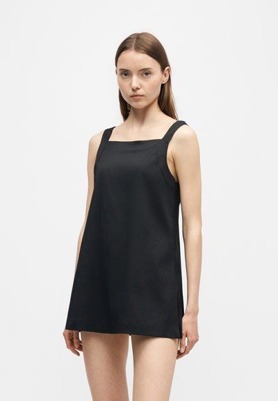 Jeune femme aux cheveux bruns et raides portant une mini-robe noire sans manches à larges bretelles sur un fond blanc uni.