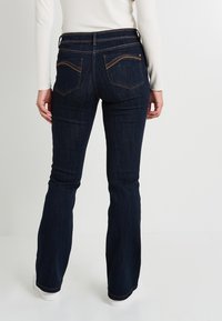 Mujer vestida con jeans de bota ancha de color azul oscuro y una blusa de manga larga blanca, de pie con las manos relajadas a los lados frente a un fondo sencillo.