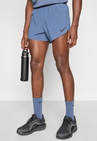 Μπλε αθλητικά σορτς από ελαφρύ υλικό, με το μαύρο λογότυπο της Nike. Μαύρα αθλητικά παπούτσια με γκρι λεπτομέρειες και μπλε ριγέ κάλτσες.