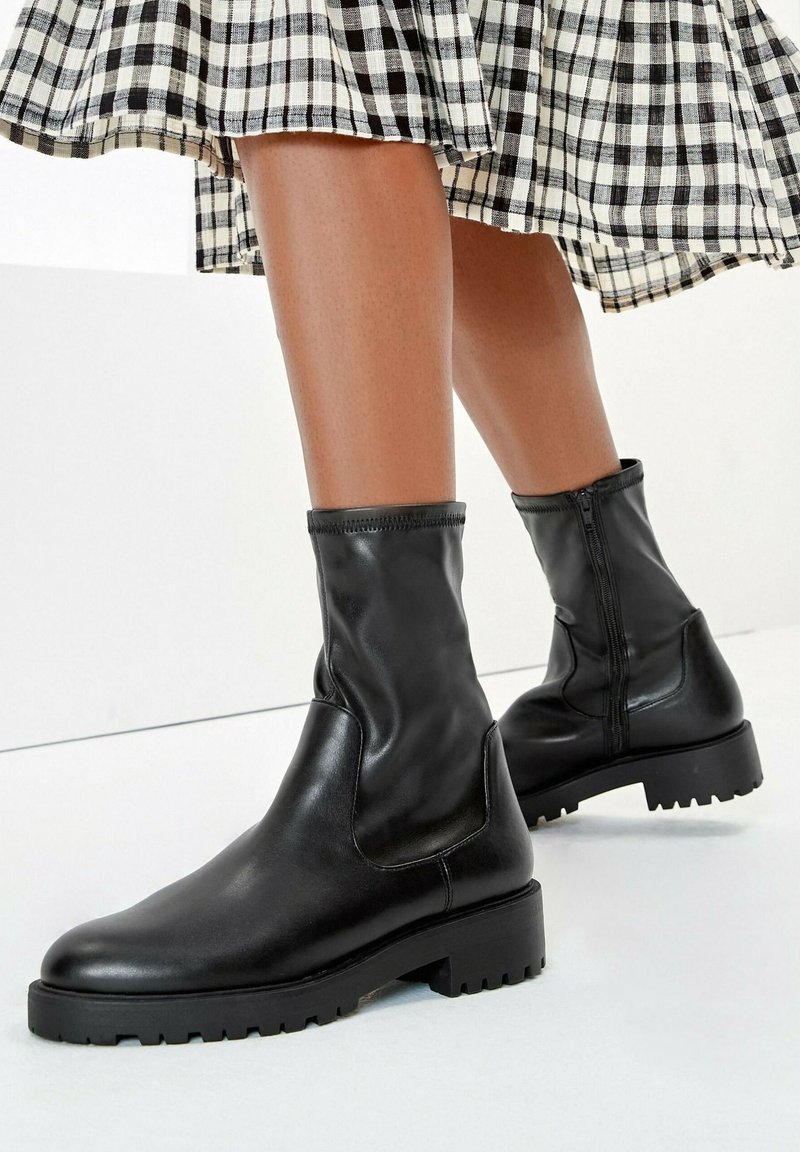 Jambes portant des bottes en cuir noir mi-mollet avec des semelles épaisses et des fermetures éclair latérales, associées à une jupe à carreaux noir et blanc.