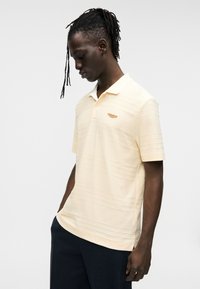 Πόλο μπλούζα σε ανοιχτό κίτρινο ύφασμα, με υφασμάτινο ριγωτό σχέδιο και μικρό λογότυπο στο αριστερό στήθος. Κοντά μανίκια, κλασικό γιακά.