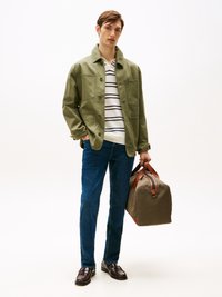 Casaco de algodão verde com múltiplos bolsos, polo às riscas brancas, jeans de ganga azuis e mocassins castanhos. Segura uma mala de lona verde-oliva com accentos castanhos.