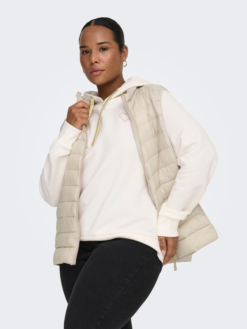 Ljusbeige vadderad väst med quiltat mönster, kombinerad med en vit hoodie med ett hjärtdesign med gulddetaljer, över svarta jeans.