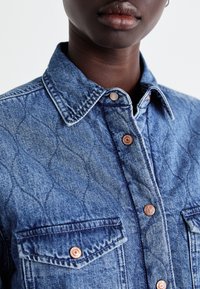 Tums zils denima krekls ar kvadrātveida faktūru, kam ir norādīta apkakle, vara pogas un divas krūšu kabatas. Tīras šuves.