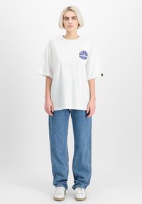 Biela voľná veľká tričko s fialovým potlačou "ALPHA INDUSTRIES" na prednej strane, spárované s modrými voľnými džínsami a bielymi teniskami.