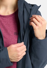 Giacca nera con tessuto testurizzato, zip frontale e foderato in rete; indossata sopra una t-shirt corta maroon con un dettaglio del logo.