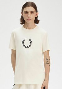 Camiseta corta de manga con tejido suave en color crema, que presenta un diseño de corona de laurel bordada en negro en el pecho. Cuello redondo sencillo.