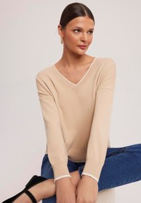 Beżowy sweter dzianinowy z dekoltem w serek, z długimi rękawami i białymi wykończeniami, w połączeniu z niebieskimi dżinsami. Miękka faktura i luźny krój.
