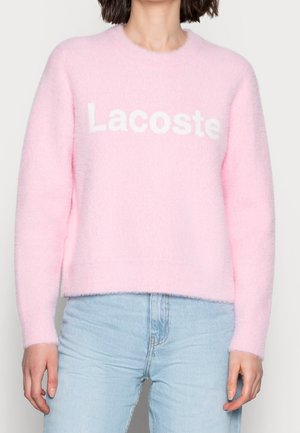 Personne portant un pull doux rose pâle avec le texte "Lacoste" en blanc, associé à un jean bleu clair sur un fond uni.