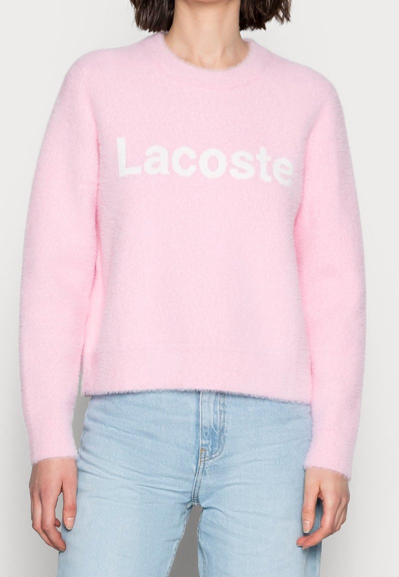 Personne portant un pull doux rose pâle avec le texte "Lacoste" en blanc, associé à un jean bleu clair sur un fond uni.