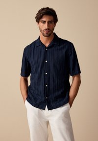 Marineblå kortærmet skjorte med knapper, vertikal ribbet struktur og klassisk krave, kombineret med hvide bukser, foran en neutral baggrund.