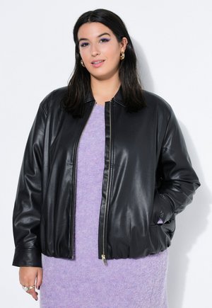 Kvinde med mørkt hår iført guldfarvede øreringe, sort læderjakke og lyslilla strikket kjole, stående mod hvid baggrund.