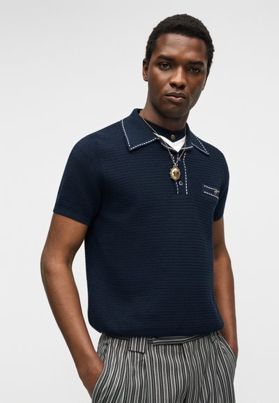 Homme portant un polo bleu marine texturé à manches courtes avec des surpiqûres blanches, un collier pendentif en or et un pantalon rayé gris, mains dans les poches, expression neutre.